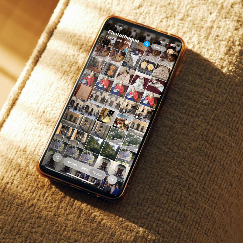 Téléphone posé sur un canapé ensoleillé, affichant une photothèque remplie de photos de famille.